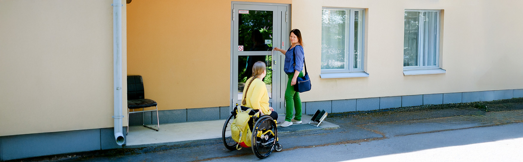 Nainen lähestyy pyörätuolissa talon ovea, jossa toinen nainen seisoo.